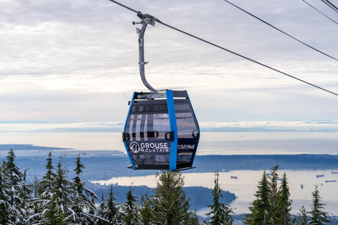 Blue Grouse Gondola ascending Grouse Mountain with Burrard Inlet and the Vancouver skyline visible in the background, under clear skies. Image courtesy of Grouse Mountain